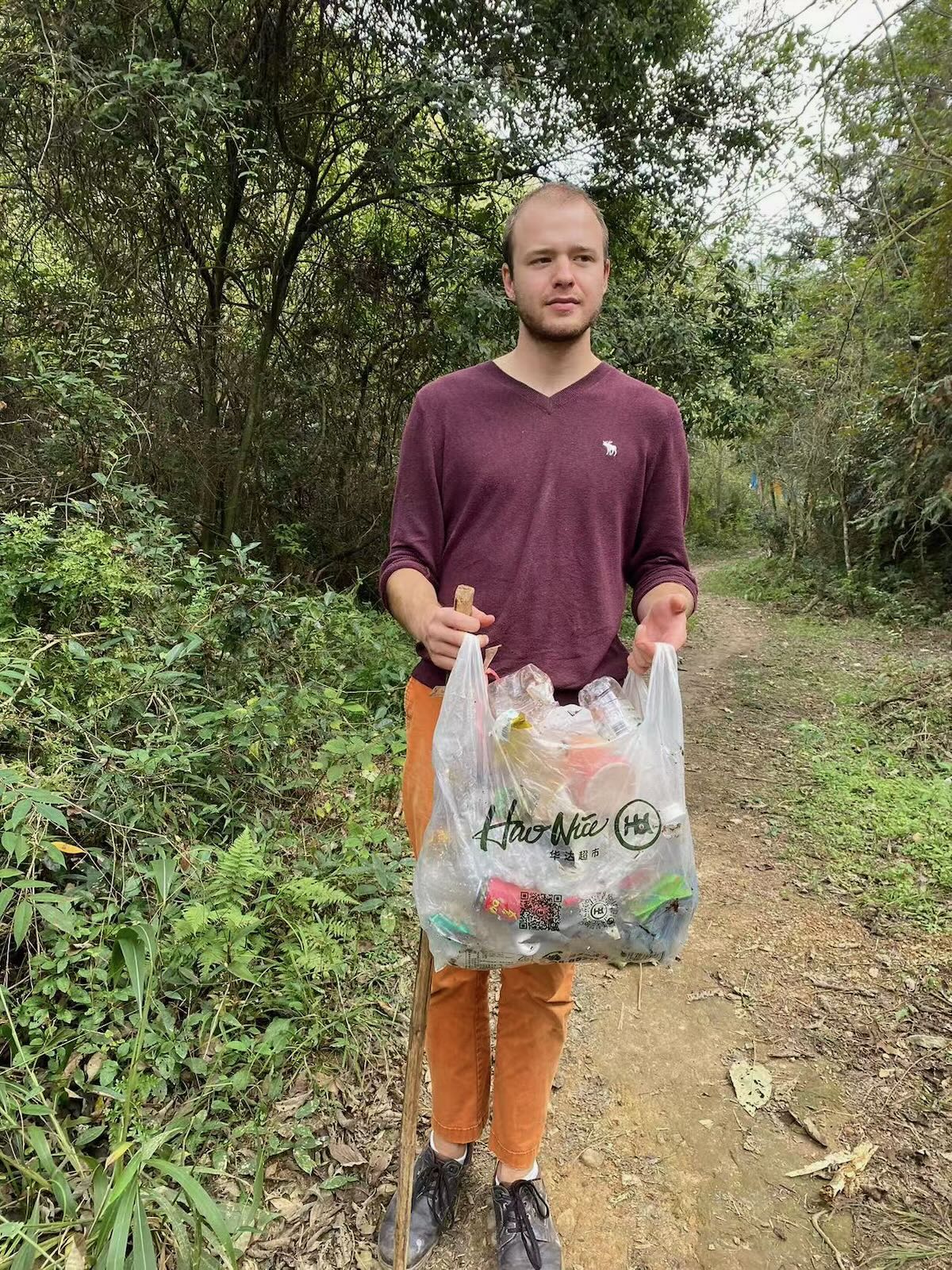 a tall blond guy looking a bit confused and holding a plastic bag full of trash items.